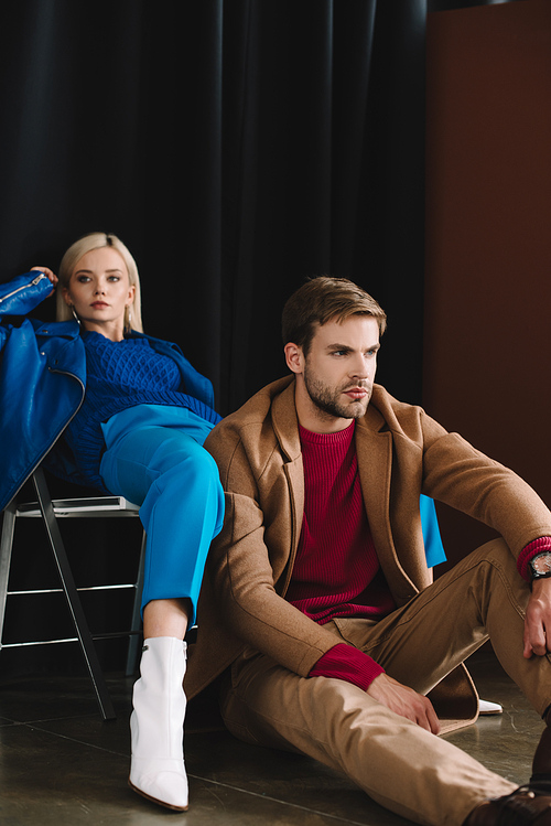 stylish blonde girl in blue leather jacket sitting on chair near man in brown coat