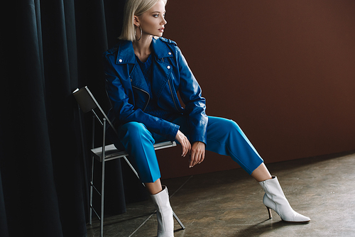 attractive blonde woman wearing leather jacket and heels sitting on chair on brown