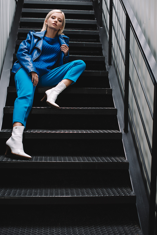 blonde woman wearing blue leather jacket and heels sitting on stairs