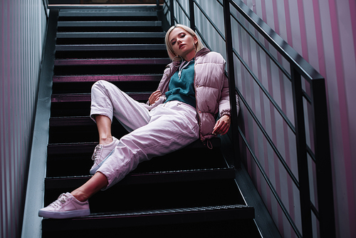 confident blonde girl in down jacket and sneakers sitting on stairs