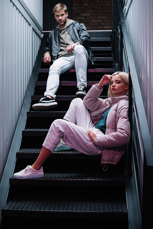 stylish young man and woman in casual autumn outfits sitting on stairs and 