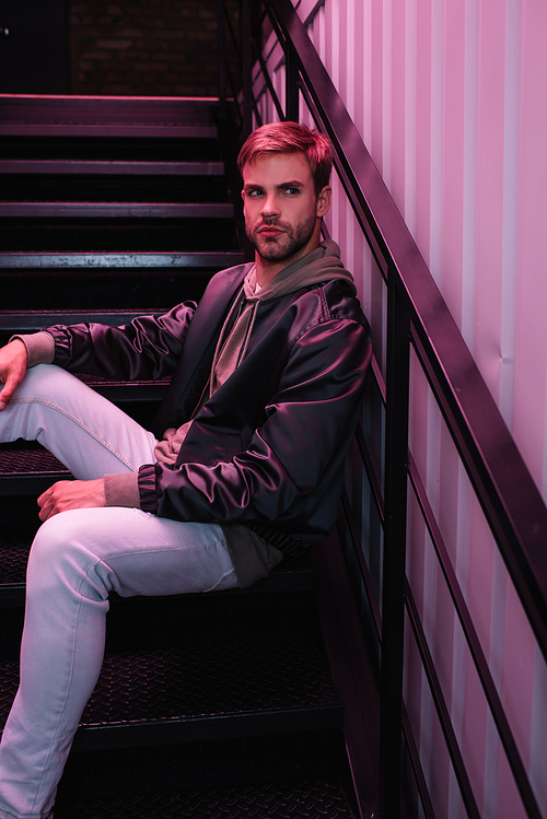 pensive bearded young man in jacket sitting on stairs