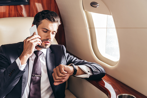 handsome businessman in suit talking on smartphone and looking at wristwatch in private plane