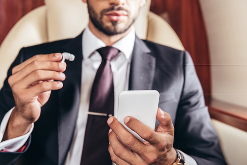 cropped view of businessman holding earphone and using smartphone in private plane