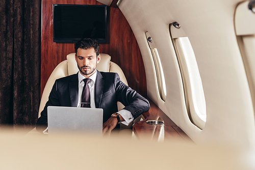 selective focus of handsome businessman in suit using laptop in private plane