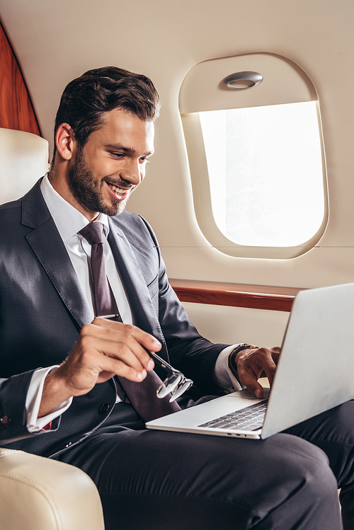 smiling businessman in suit using laptop and holding glasses in private plane