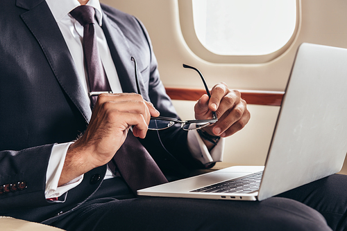 cropped view of businessman in suit with laptop holding glasses in private plane