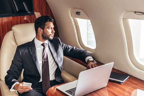 handsome businessman in suit looking through window and holding smartphone in private plane