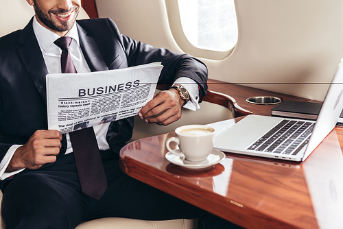 cropped view of smiling businessman in suit reading business newspaper in private plane