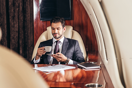 selective focus of handsome businessman in suit drinking coffee in private plane