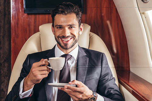 handsome businessman in suit smiling and holding cup of coffee in private plane