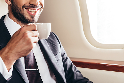 cropped view of smiling businessman in suit holding cup of coffee in private plane