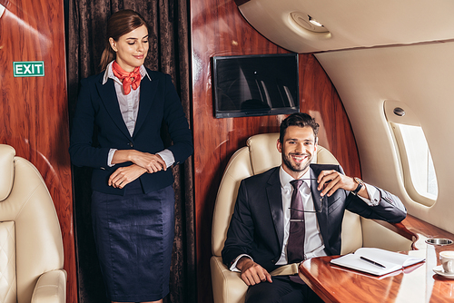 flight attendant in uniform looking at smiling businessman in private plane