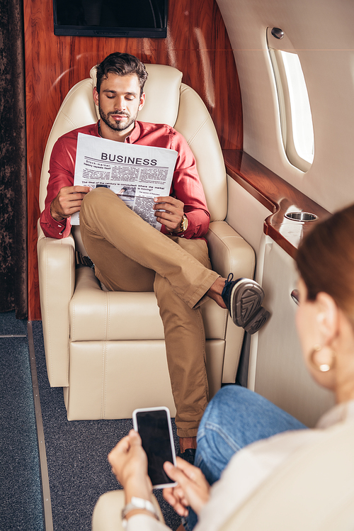 selective focus of boyfriend reading newspaper and girlfriend holding smartphone in private plane