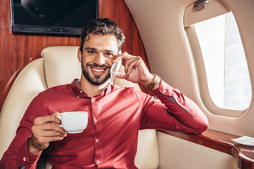 handsome man in shirt talking on smartphone and holding cup in private plane