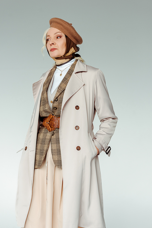 Beautiful senior woman in trench coat and beret looking away isolated on grey