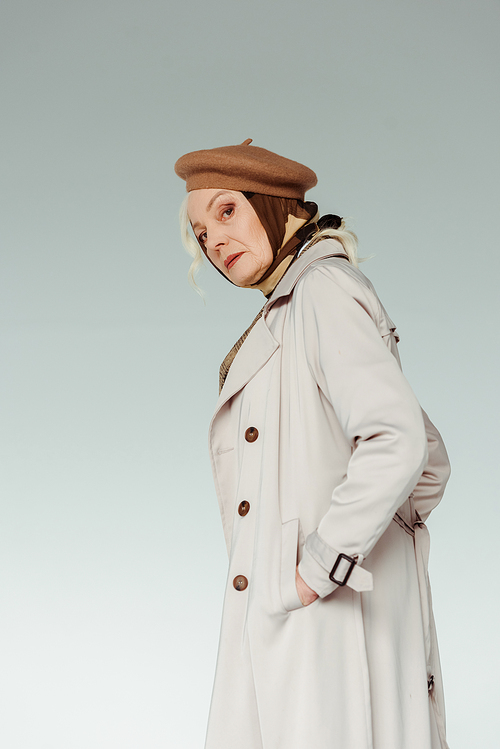 Side view of stylish senior woman  while holding hand in pocket of coat isolated on grey