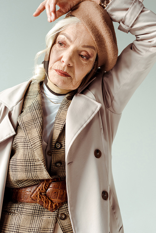 Elegant elderly woman in trench coat and beret posing isolated on grey