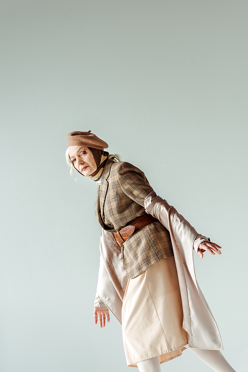 Side view of stylish senior woman in trench coat and beret  isolated on grey