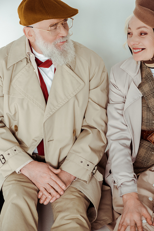 Stylish senior couple smiling at each other while sitting on white surface on grey background