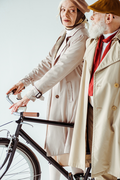 Side view of stylish elderly woman with bicycle looking at elegant man in trench coat isolated on white
