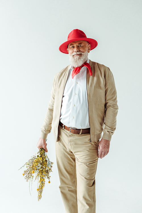 Fashionable elderly man in red hat holding wildflowers and smiling at camera isolated on white