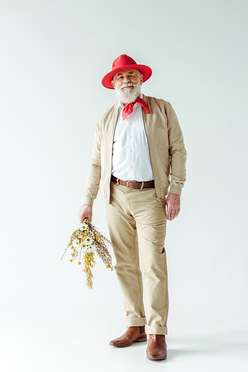 Full length of stylish senior man in red hat smiling at camera and holding wildflowers on white background