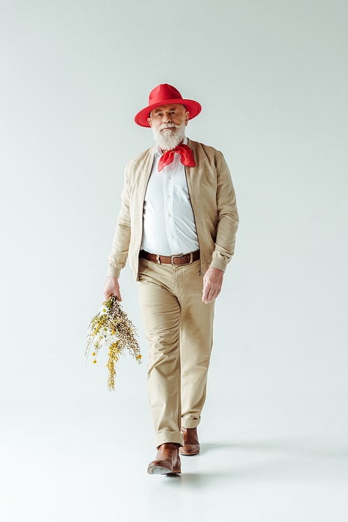 Full length of handsome senior man in red hat holding bouquet of wildflowers while walking on white background