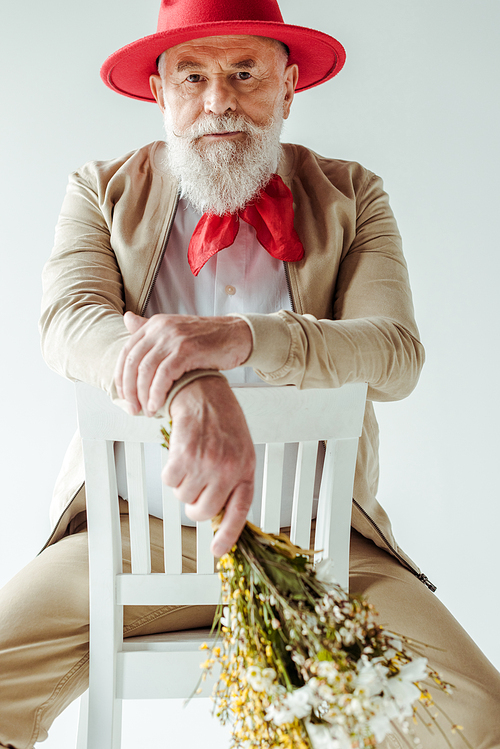 Selective focus of stylish senior man in red hat holding wildflowers and  on chair isolated on white