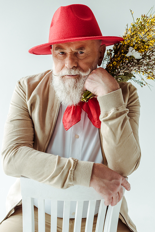 Handsome senior man in red hat holding wildflowers and  on chair isolated on white