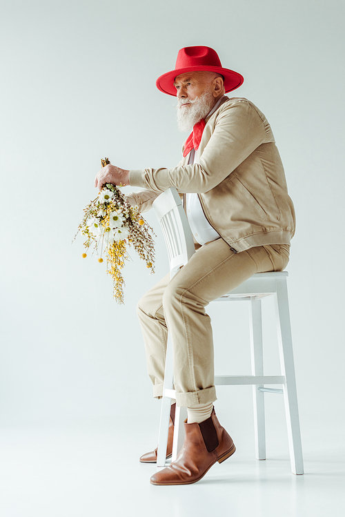 Side view of handsome senior man in red hat holding wildflowers while sitting on chair on white background