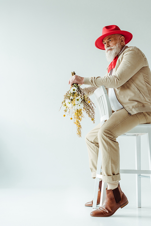 Full length of stylish elderly man in red hat holding bouquet of wildflowers while sitting on chair on white background
