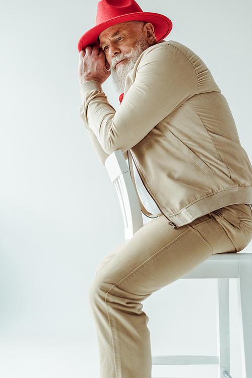 Side view of stylish senior man in red hat sitting on chair isolated on white