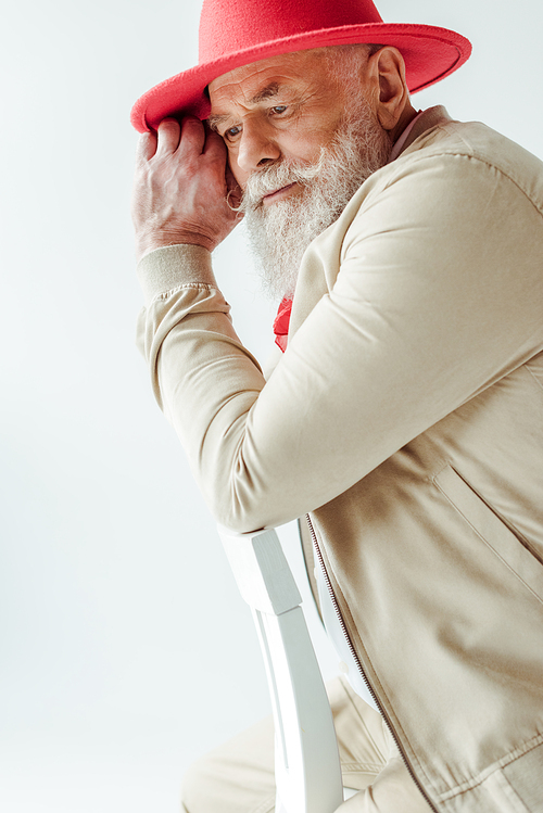 Side view of handsome elderly man in red hat looking away on chair isolated on white