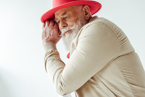 Side view of stylish senior man in red hat looking away while sitting on chair isolated on white