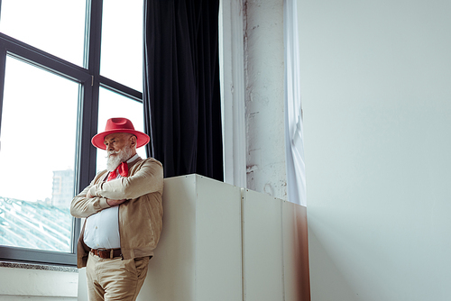 Stylish senior man in red hat standing near window in photo studio