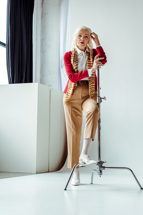 Elegant senior woman standing near tripod on white background in photo studio