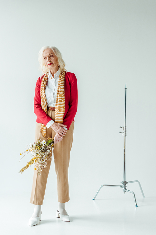 Full length of beautiful senior woman holding bouquet of wildflowers near tripod on white background