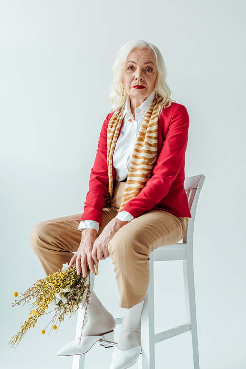 Stylish senior woman  while holding wildflowers on chair isolated on white