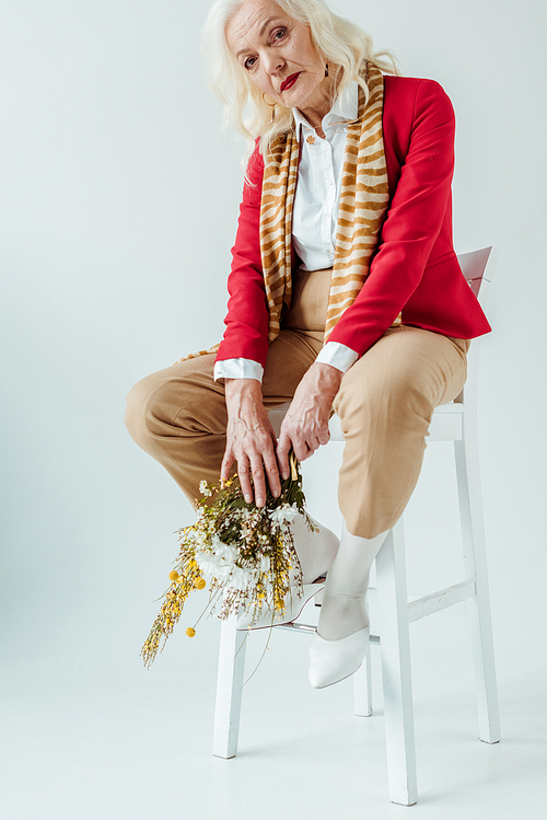 Fashionable elderly woman sitting on chair and holding bouquet of wildflowers on white background