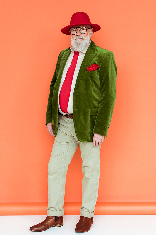 Full length of handsome senior man in hat and eyeglasses  on white surface on coral background