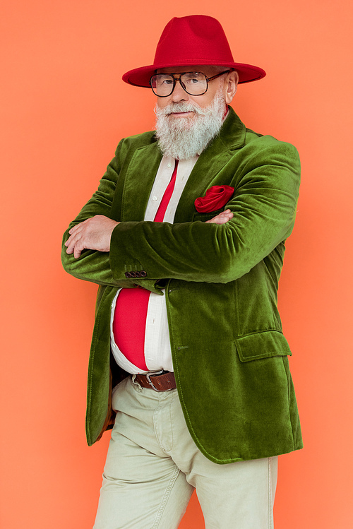 Stylish senior man in eyeglasses and hat with crosses arms  isolated on coral