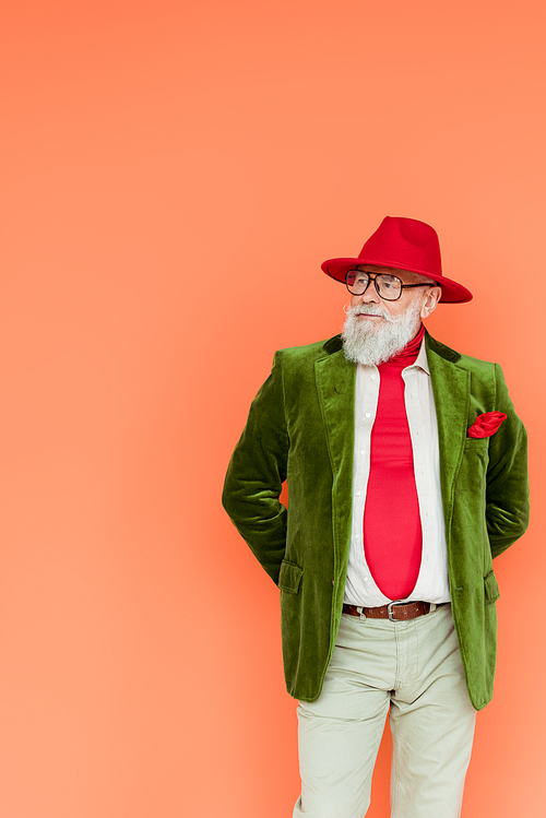 Fashionable elderly man in hat and eyeglasses looking away isolated on coral