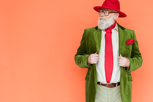 Handsome senior man in hat holding jacket and looking away isolated on coral with copy space