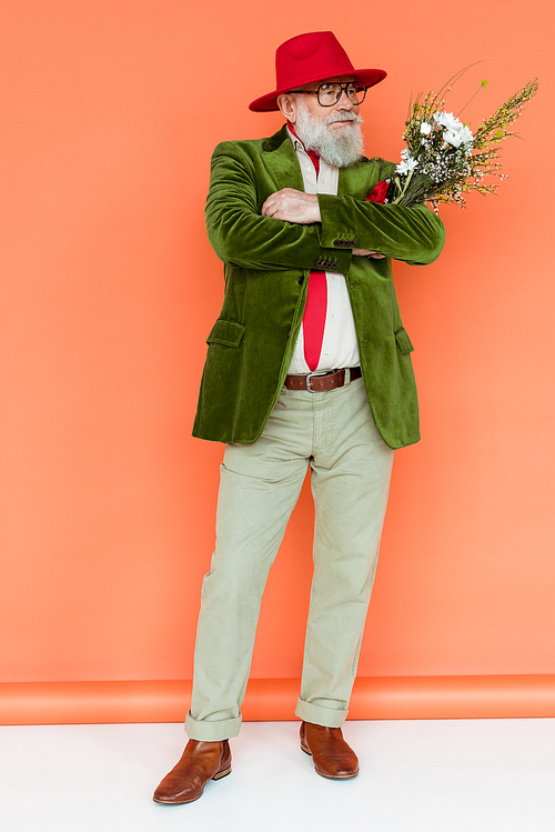 Full length of handsome elderly man with crossed arms holding wildflowers on white surface on coral background