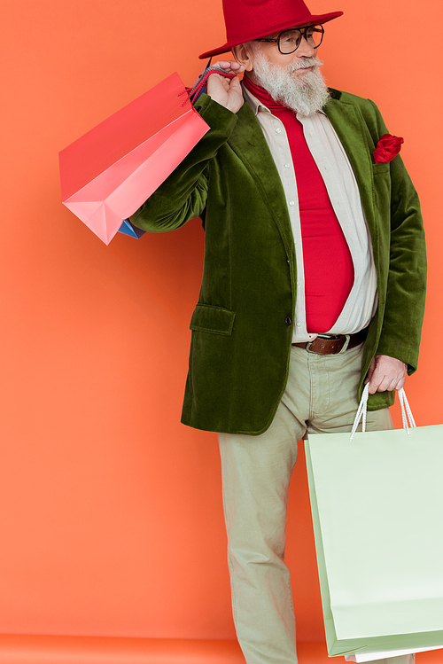 Fashionable elderly man holding shopping bags and looking away on coral background