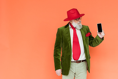 Handsome senior man in hat and eyeglasses holding smartphone with blank screen isolated on coral