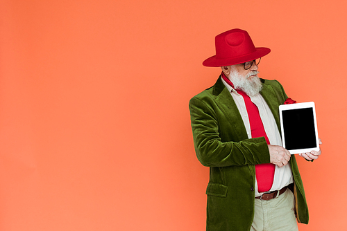 Handsome elderly man in hat and eyeglasses holding digital tablet isolated on coral with copy space