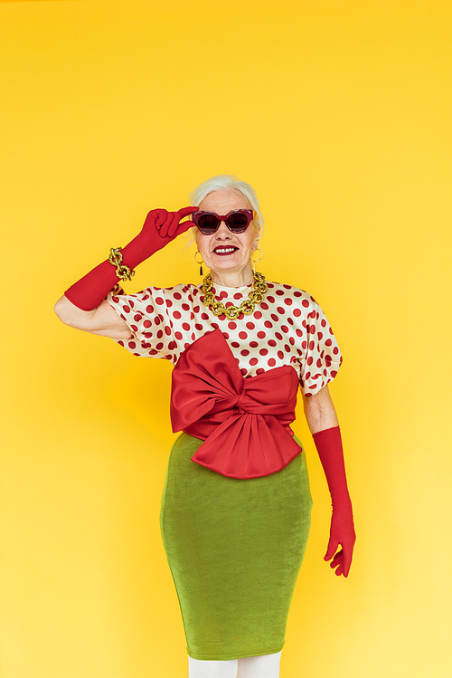 Stylish senior woman smiling and holding sunglasses isolated on yellow