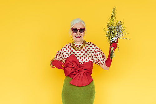 Stylish senior woman smiling while holding bouquet of wildflowers isolated on yellow